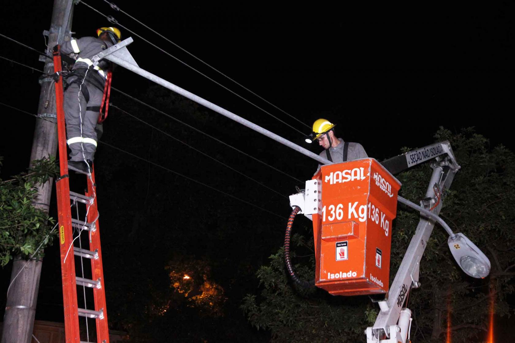 Homens fazem manutenção e troca de luz. Um deles está em uma escada encostada em um poste. O outro, está dentro de um cesto de caminhão com braço basculante. É noite. #Pracegover