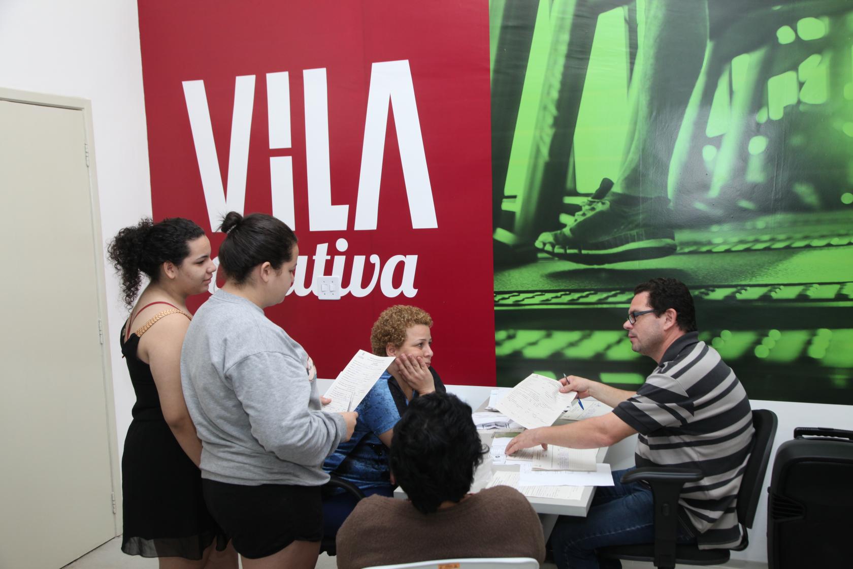 Em uma sala, homem está sentado à frente de uma mesa com papéis. Há duas mulheres sentadas e outras duas em pé. Ao fundo há um painel onde se lê Vila Criativa. #Pracegover