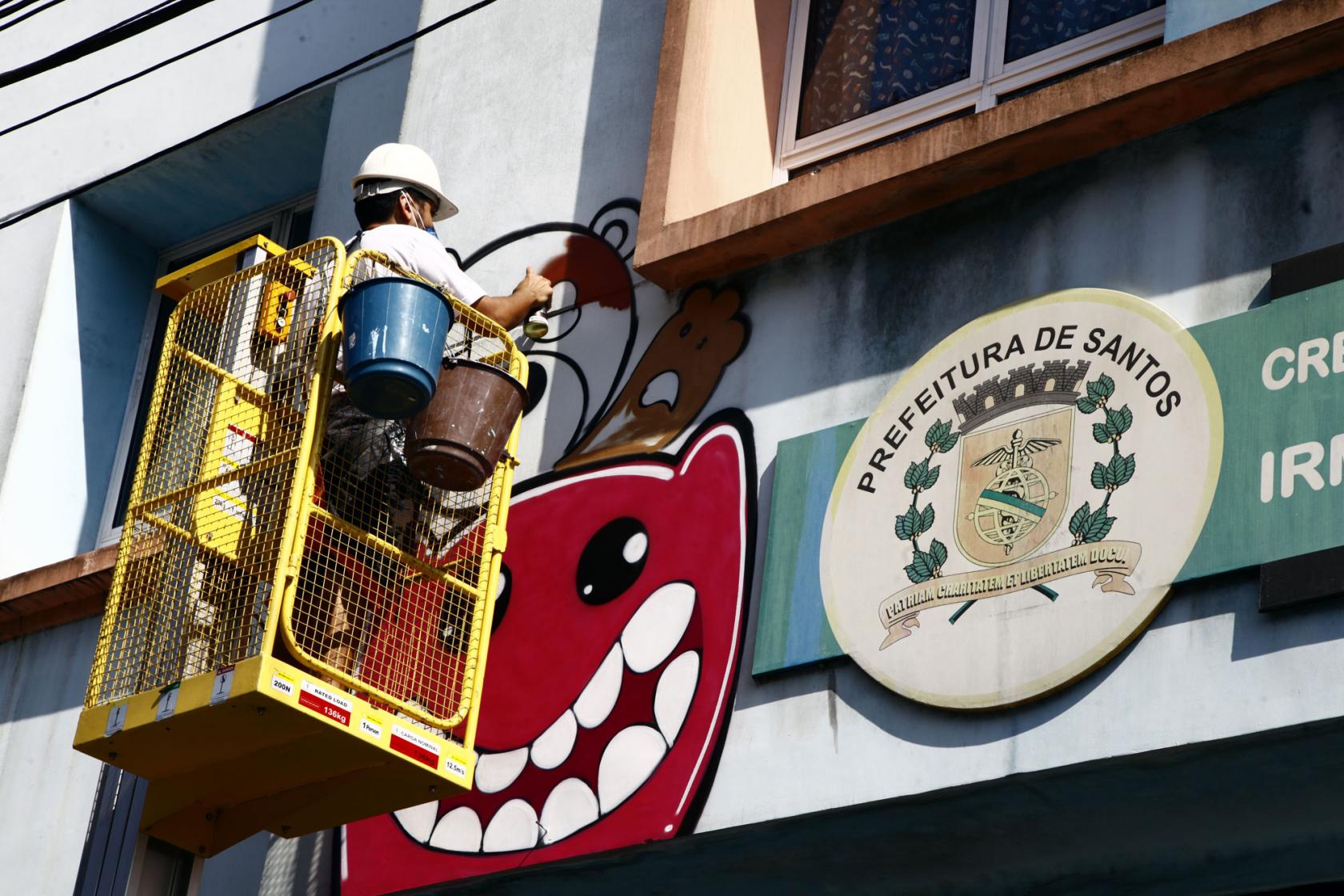 Grafiteiro está dentro de plataforma elevatória fazendo arte em grafite. Ao lado direito aparece o brasão de Santos na placa da escola. #Pracegover