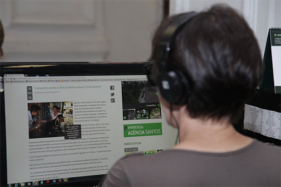 Na imagem, mulher está sentada em frente ao computador com fonte de ouvido. E na tela é possível observar foto e texto sobre a campanha de vacinação contra raiva em Santos.