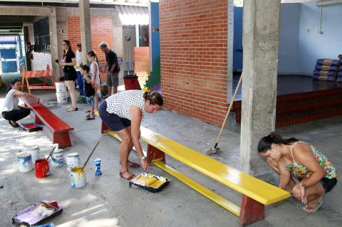 Mães de alunos e voluntários pintam bancos de madeira do pátio da escola. #pracegover