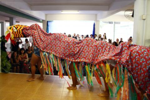 Apresentação artística dos alunos da escola Lourdes Ortiz à comitiva. Crianças estão no palco com os pés descalços e estão todas cobertas por um tecido colorido como fios também coloridos presos em toda a extensão lateral. #pracegover