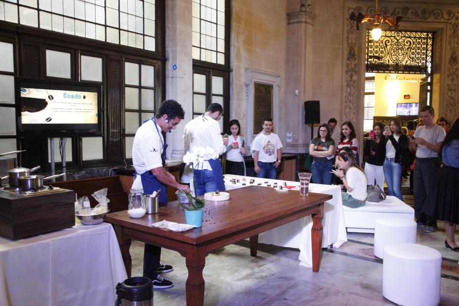 No saguão do museu há uma mesa onde estão sendo preparados pratos à base de café por im instrutor que está em pé. Há diversos recipientes de cozinha. Ao lado esquerdo dele há outro instrutor, à frente de outra mesa, também preparando alguma receita. O público está em pé atento às orientações. #Pracegover