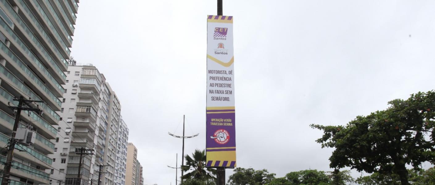 banner afixado em poste em canteiro central de avenida da orla. Carros passam nas duas pistas, indo e vindo. #paratodosverem