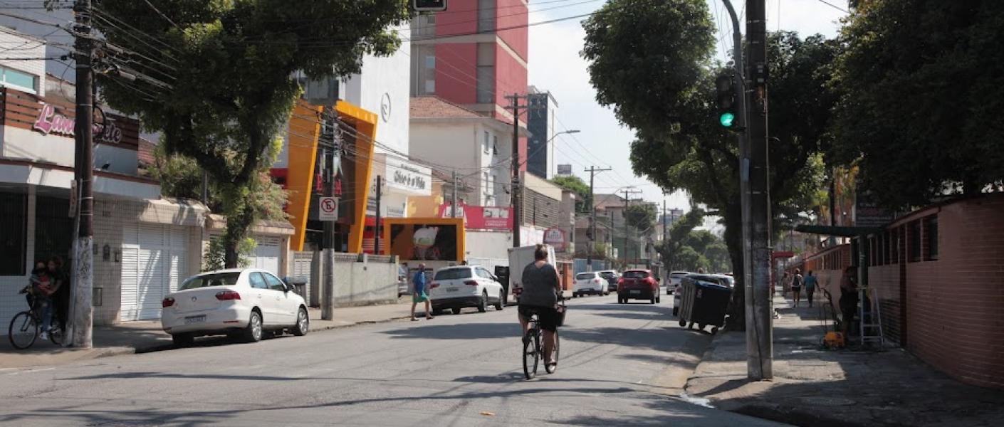 trecho de rua com calçadas, carros estacionados dos dois lados e uma mulher de bicicleta indo adiante da foto. #paratodosverem