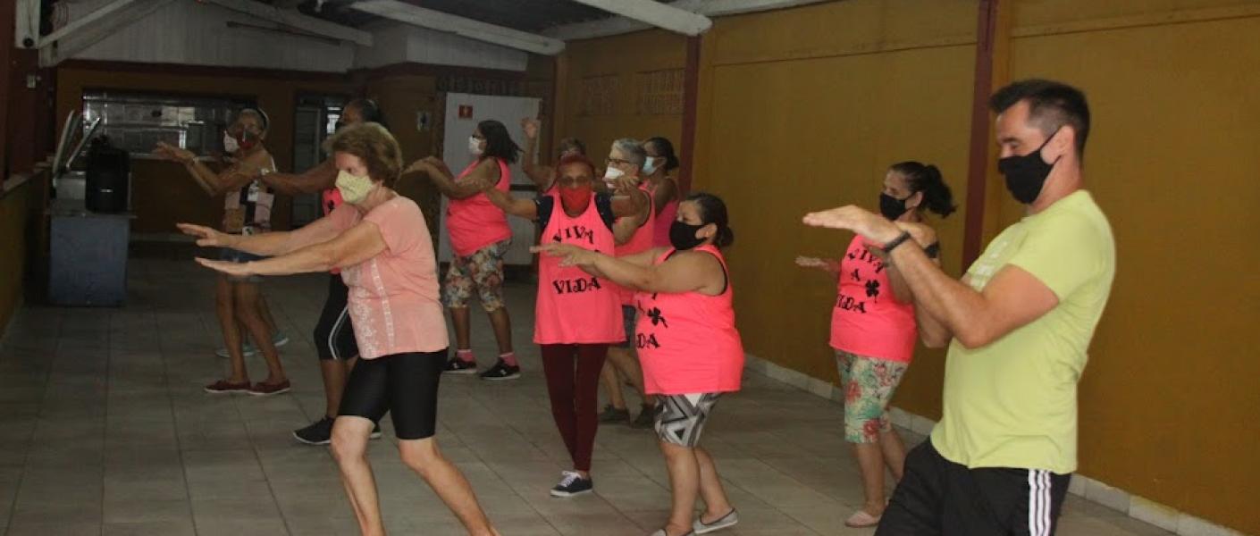 mulheres estão empé se movimentando com os braços para a frente, seguindo as orientações do professor. #paratodosverem