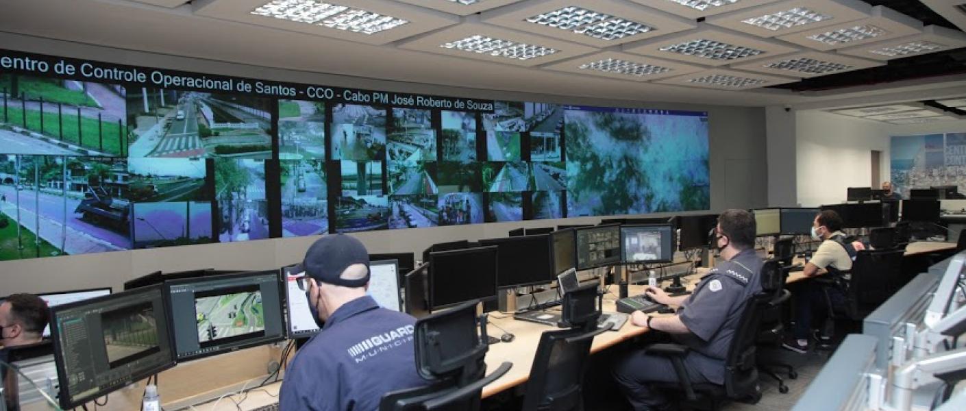 Monitoramento urbano é feito todos os dias (24 horas). Guardas e policiais observam monitores de computadores e telões. #paratodosverem