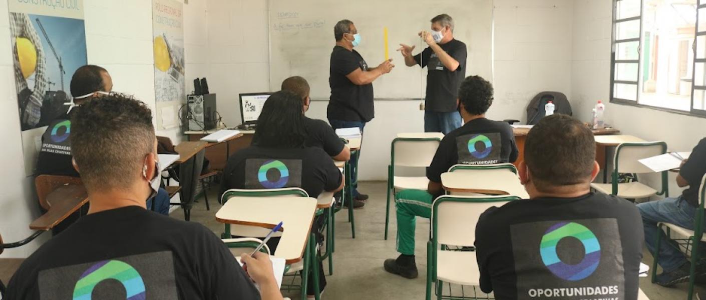 alunos estão sentados em respectivas carteiras. Eles estão de costas para a foto, atentos ao professor e mais um homem, que mexem num objeto. #paratodosverem