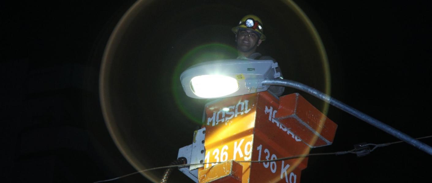 homem está em cesto no alto de poste mexendo em luminária acesa. #paratodosverem