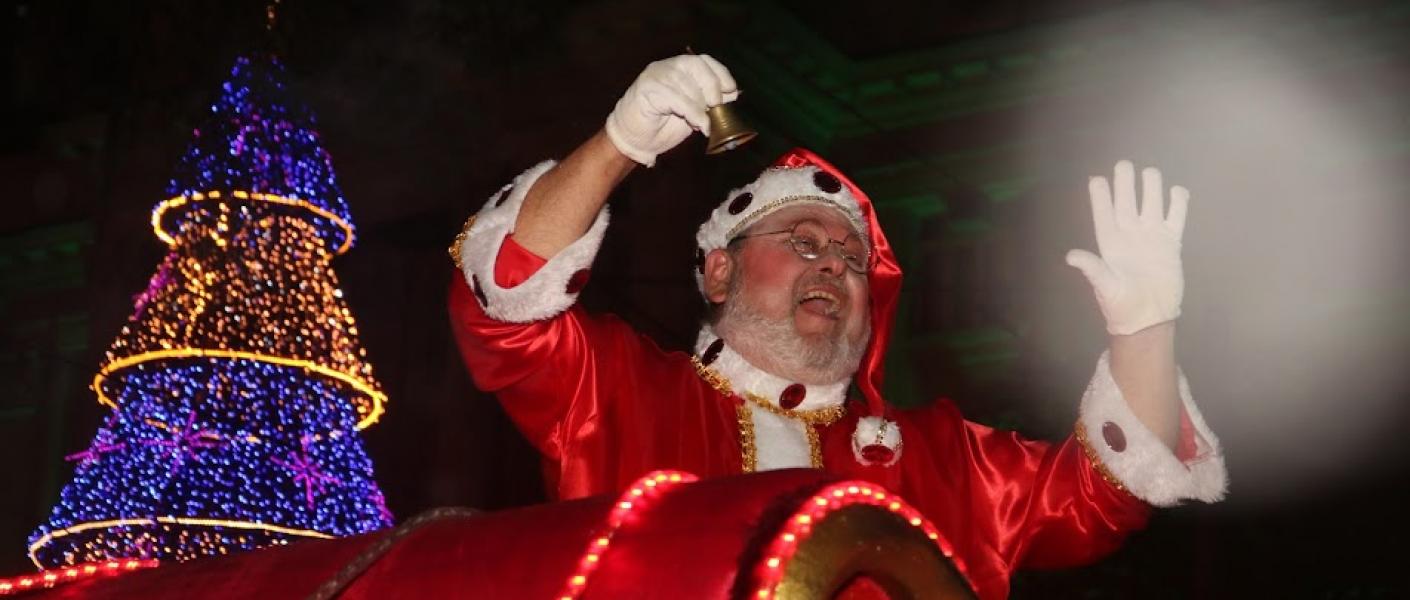 papai noel passa de treno diante de grande árvore de natal iluminada. Ele toca o sino e saúda o público (que não aparece na imagem). Ao fundo, o paço municipal. #paratodosverem