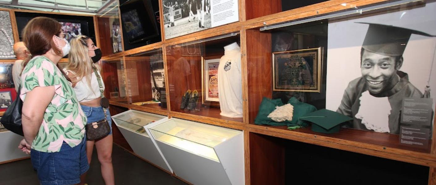 área interna do museu com galeria de imagens. Em primeiro plano, à direito, uma foto de pelé com beca e capelo. Pessoas estão olhando. #paratodosverem