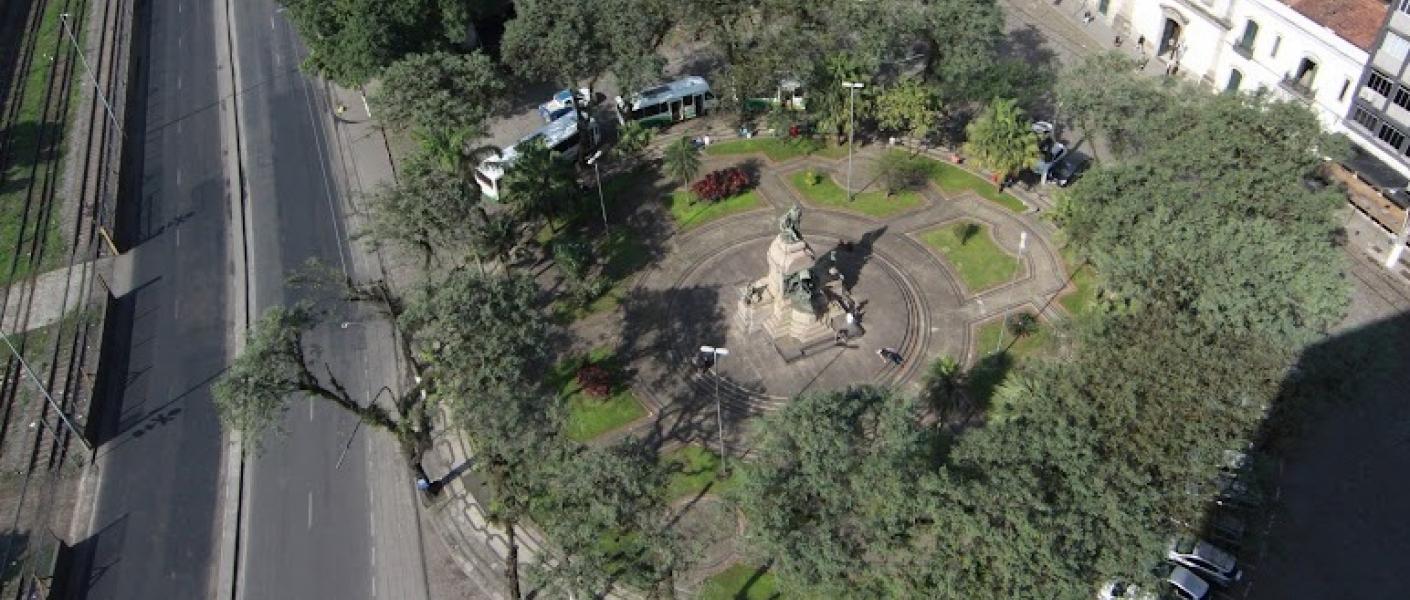 vista aérea da praça com monumento ao centro. A praça é rodeada por árvores. #paratodosverem