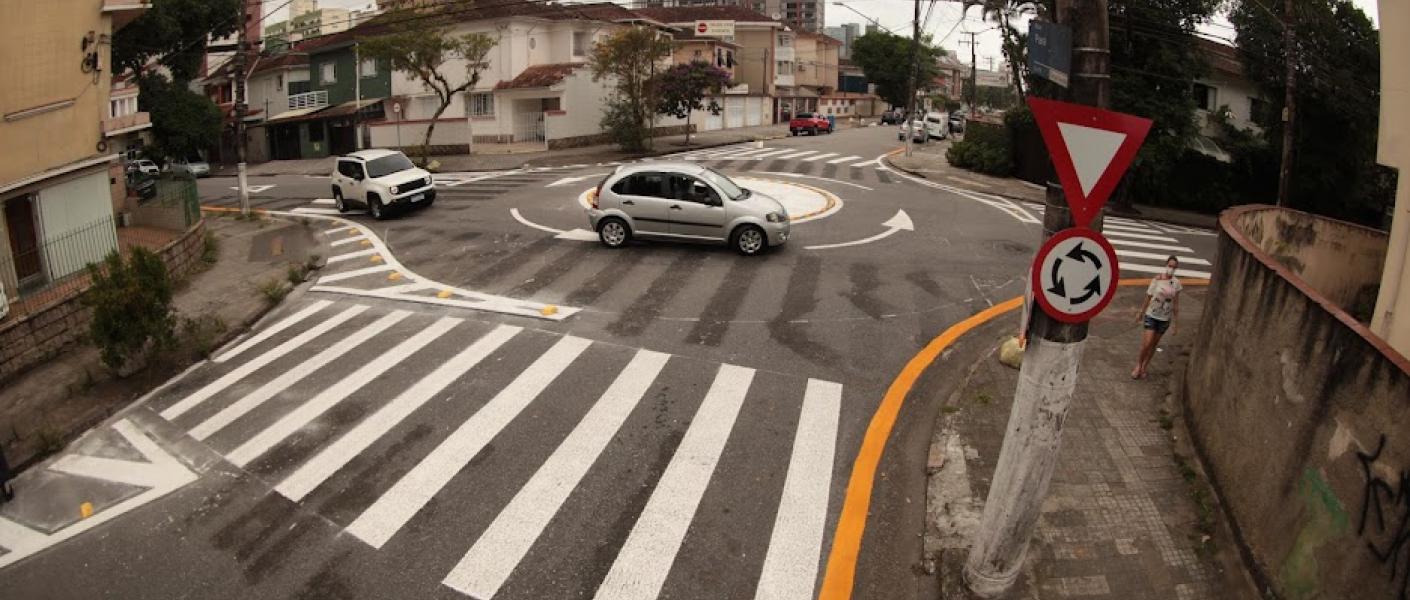 Carros passam por rotatória enquanto pedestre anda pela calçada. #pratodosverem