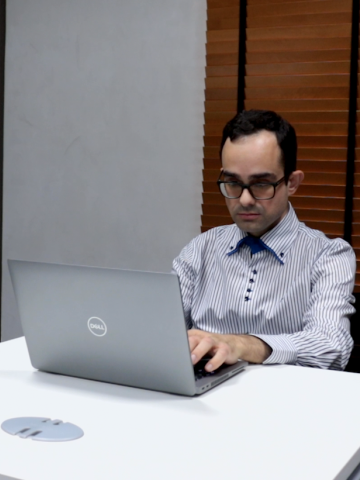 Rapaz de óculos e camisa social trabalha sentado diante de laptop. #pratodosverem
