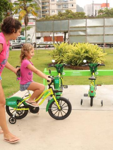 criança pedala bicicleta com a mãe em pé ao lado. #paratodosverem
