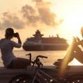 duas pessoas estão sentadas na mureta da orla assistindo à saída de um navio. Os dois fotografam com celulares. A foto foi feita ao por do sol, que cai e brilha do lado direito. #paratodosverem