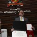 homem está em pé segurando uma caixa com medalha e uma pasta com um certificado. #paratodosverem