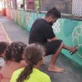 artista desenha e pinta bola no muro de quadra. Crianças, de costas para a foto, observam os movimentos do artista. #paratodosverem