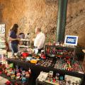 feira de artesanato com mesas cheias de pequenas peças. Duas pessoas estão em pé em torno dessas mesas. #paratodosverem 