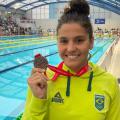 Beatriz Dizotti posa para foto segurando medalha e de costas para uma piscina. #paratodosverem