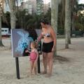Mulher e criança observam placa educativa instalada na faixa de areia da Praia do José Menino. #pratodosverem