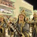 Integrante de bateria movimenta rocar chocalho. Sobre o adereço em sua cabeça se lê: Fé. Ela sorri para a foto Em volta há vários músicos. Ao fundo está a plateia. #Paratodosverem