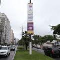 banner afixado em poste em canteiro central de avenida da orla. Carros passam nas duas pistas, indo e vindo. #paratodosverem