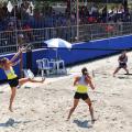 duas equipes jogam beach tennis, com duas duplas em lados opostos da rede ao centro da quadra de areia. São mulheres. Elas seguram raquetes e fazem movimentos do jovo. Ao lado esquerdo, a arquibancada. #paratodosverem