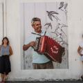 Beatriz e Carolina estão em pé ao lado da imagem de um homem tocando concertina. #Paratodosverem