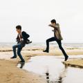 Dois jovens pulando uma faixa de água na areia da praia #paratodosverem