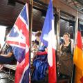 Bandeiras de diversas nações estão na lateral de um bonde. Mulheres seguram os pavilhões. #Pracegover