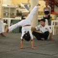 menina está dando estrela em aula de capoeira. Suas pernas estão abertas no ar. As duas mãos no chão. Ao fundo, mais crianças agachadas. 