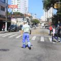 agente está no meio de uma rua, de costas para a foto e de frente para faixa de pedestres. Algumas pessoas cruzam a rua. #paratodosverem