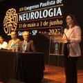 palco de evento com a vice prefeita em pé falando. cinco pessoas estão sentadas a uma mesa. #paratodosverem 