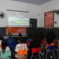 jovens estão sentados em cadeiras, de costas para a foto, assistindo a uma projeção num telão na parede. #paratodosverem