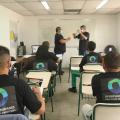 alunos estão sentados em respectivas carteiras. Eles estão de costas para a foto, atentos ao professor e mais um homem, que mexem num objeto. #paratodosverem