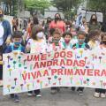 Crianças caminham pela rua, acompanhadas por professoras. Elas carregam uma faixa onde se lê UME DOS ANDRADAS VIVA A PRIMAVERA. #paratodosverem