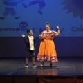 Personagem está no palco ao lado de menino usando uniforme escolar. #paratodosverem