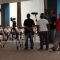 equipe de produção visual, com câmeras está dinate de de crianças que estão sentadas em cadeiras dispostas lado a lado num grande salão. #paratodosverem