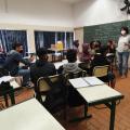 sala de aula com alunos sentados e duas mulheres em pé. Uma delas fala ao grupo. #paratodosverem