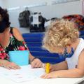 Educadora está ao lado de criança que desenha sobre uma mesa. #paratodosverem