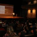 Servidores sentados na plateia do teatro assistem exibição de secretário, em pé no palco, abaixo de telão. #pratodosverem