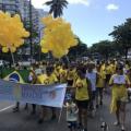 pessoas caminhando na avenida, usando roupas amarelas. Algumas seguram balões amarelos em alusão à endometriose. Duas pessoas à frente da marcha seguram uma faixa onde se lê Endomarcha. #paratodosverem