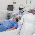 Dois profissionais de saúde uniformizados estão atendendo um paciente no leito de hospital. #paratodosverem