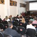 Pessoas sentadas em um auditório observam pessoa falando e telão ao fundo #paratodosverem