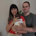 casal posa para foto. A mulher segura o bebê. Com parte do corpo atrás da mulher, homem coloca a mão sobre o corpinho do bebê. #paratodosverem