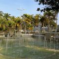 Vista geral de praça com chafariz e palmeiras ao fundo. #paratodosverem 