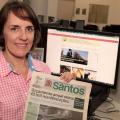 Márcia está segurando uma antiga edição do Diário Oficial em papel. Atrás dela, um monitor de desktop está aberto no  Santos Portal. #Pracegover