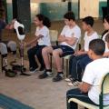 Pauê está sentado em uma cadeira usando e mostra exemplar de próteses para os alunos. Uma das meninas, ao lado dele, toca na peça. Os outros estudantes observam. #Pracegover