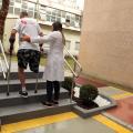 mulher usando avental branco auxilia homem com prótese na perna direita a subir degraus. #paratodosverem